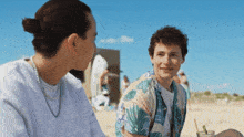 two young men are sitting on the beach and talking