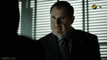 a man in a suit and tie talks to another man in front of a window
