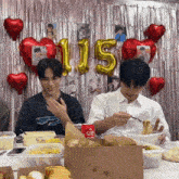 two young men are eating food in front of a wall with balloons and a number 115 balloon