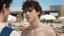 a shirtless man with curly hair talks to another shirtless man on a beach