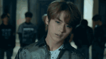 a young man wearing a jacket and a white shirt looks at the camera with a group of people in the background