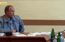 a man sits at a table with bottles of water and a subscribe button on the wall behind him