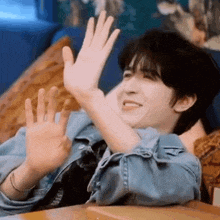 a man in a denim jacket is sitting on a couch with his hands in the air .