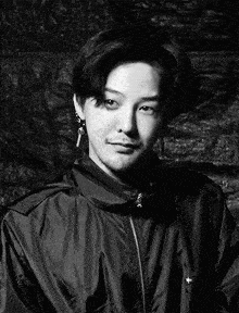 a black and white photo of a young man with a beard wearing a jacket and earrings .