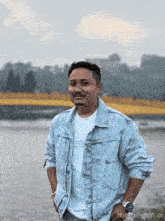 a man in a denim jacket is standing in front of a river