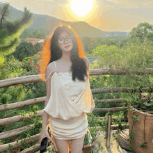 a woman wearing glasses and a white dress stands on a balcony with the sun behind her