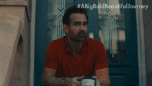 a man in a red shirt sits on a porch holding a coffee mug with #abigbold beautifuljourney written on the bottom