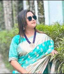 a woman wearing a blue saree and sunglasses is standing next to a bush .