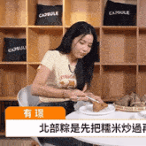 a woman sits at a table with a plate of food in front of a shelf with capsule pillows on it