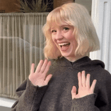 a woman with blonde hair wearing a grey sweater is smiling and waving her hands
