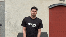 a man wearing an agon by aoc t-shirt stands in front of a red door