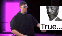 a man stands in front of a purple wall with a picture of a man behind him that says true