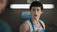 a young man with curly hair is wearing a white tank top and a blue apron
