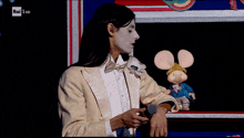 a man in a white tuxedo is holding a microphone in front of a stuffed mouse on a rai hd screen