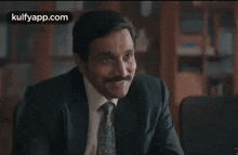 a man in a suit and tie is sitting at a desk smiling .