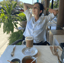 a woman sits at a table with her hands behind her head and a cup of coffee