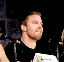 a man with a beard stands in front of microphones at comic con