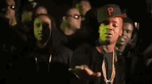 a group of men are standing in a dark room . one of the men is wearing a baseball cap with the letter p on it .