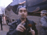 a man in a leather jacket is holding a red flower in front of a bus