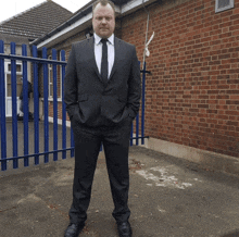 a man in a suit stands in front of a blue fence