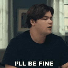 a man in a black shirt is saying `` i 'll be fine '' while sitting at a table .