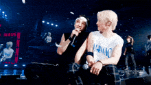 two men are sitting next to each other and one has a shirt that says maniac on it