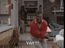 a man in a red sweater is dancing in a living room with the words `` yay '' .
