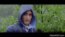 a man wearing a blue raincoat with a hood is standing in the rain next to a tree .