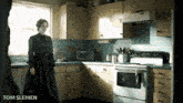 a woman in a black dress is standing in a kitchen with the name tom slemen on the bottom right