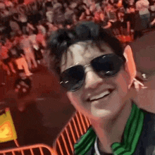 a man wearing sunglasses and a green scarf is taking a selfie in front of a crowd of people .