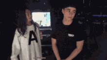 a girl wearing a white sweater with the letter a on it stands next to a boy wearing a black shirt that says text
