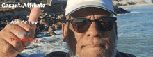 a man wearing sunglasses and a hat giving a thumbs up in front of the ocean