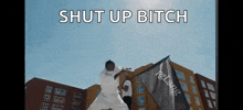 a man in a white shirt is holding a flag in front of a building and says `` shut up bitch '' .