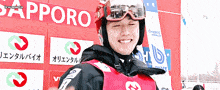 a man wearing a red helmet and goggles is smiling in front of a sign that says sapporo