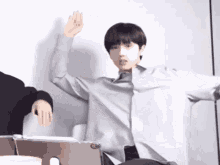 a young man in a grey shirt is sitting in front of a laptop computer and raising his hand in the air .