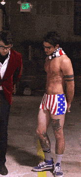 a shirtless man in american flag shorts stands next to a man in a red jacket in front of a green exit sign
