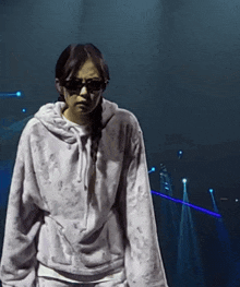 a woman wearing sunglasses and a hoodie stands in front of a blue light