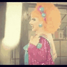 a drag queen with orange hair is wearing a pink sequined dress and earrings .