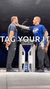 two men are standing next to each other on a stage with the words tag your it written on the bottom .