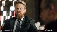 a man with a beard wearing a suit and tie is sitting in a chair with a nbc logo behind him