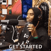 a man in a hoodie is sitting in front of a microphone holding a cup of coffee and saying get started .