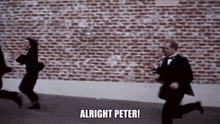 a group of people in suits and tuxedos are running down a street in front of a brick wall .
