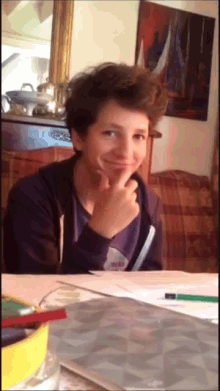 a young boy is sitting at a table with his hand on his chin and smiling
