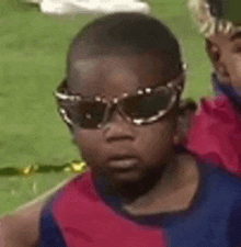 a young boy wearing sunglasses and a red and blue shirt is sitting on a soccer field .