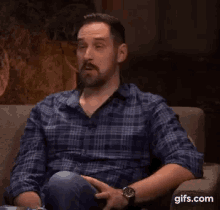 a man with a beard is sitting on a couch with a watch on his wrist and a plaid shirt on .