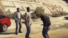 a group of men are standing in front of a wall with graffiti on it and a red car