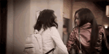 two women are standing next to each other and talking . one of the women is wearing a backpack .