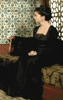 a woman in a black dress is sitting on a couch with her hands folded .
