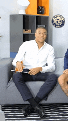 a man in a white shirt is sitting on a couch in front of a soccer logo that says mbappe live