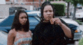 two women are standing next to each other in front of a car and one of them is eating a sandwich .